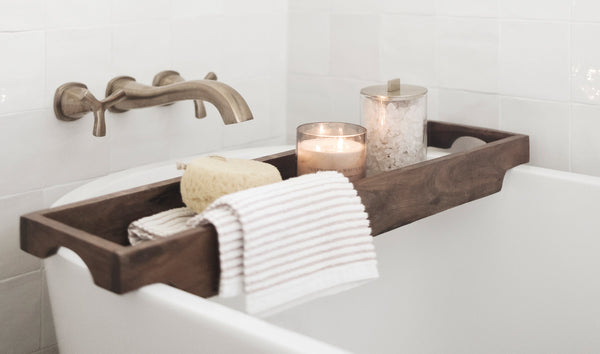 Close-up of a serene bath setup featuring a wooden bathtub tray holding a lit candle, a jar of bath salts, a soft striped towel, and a natural sponge, styled to create a calming evening ritual.