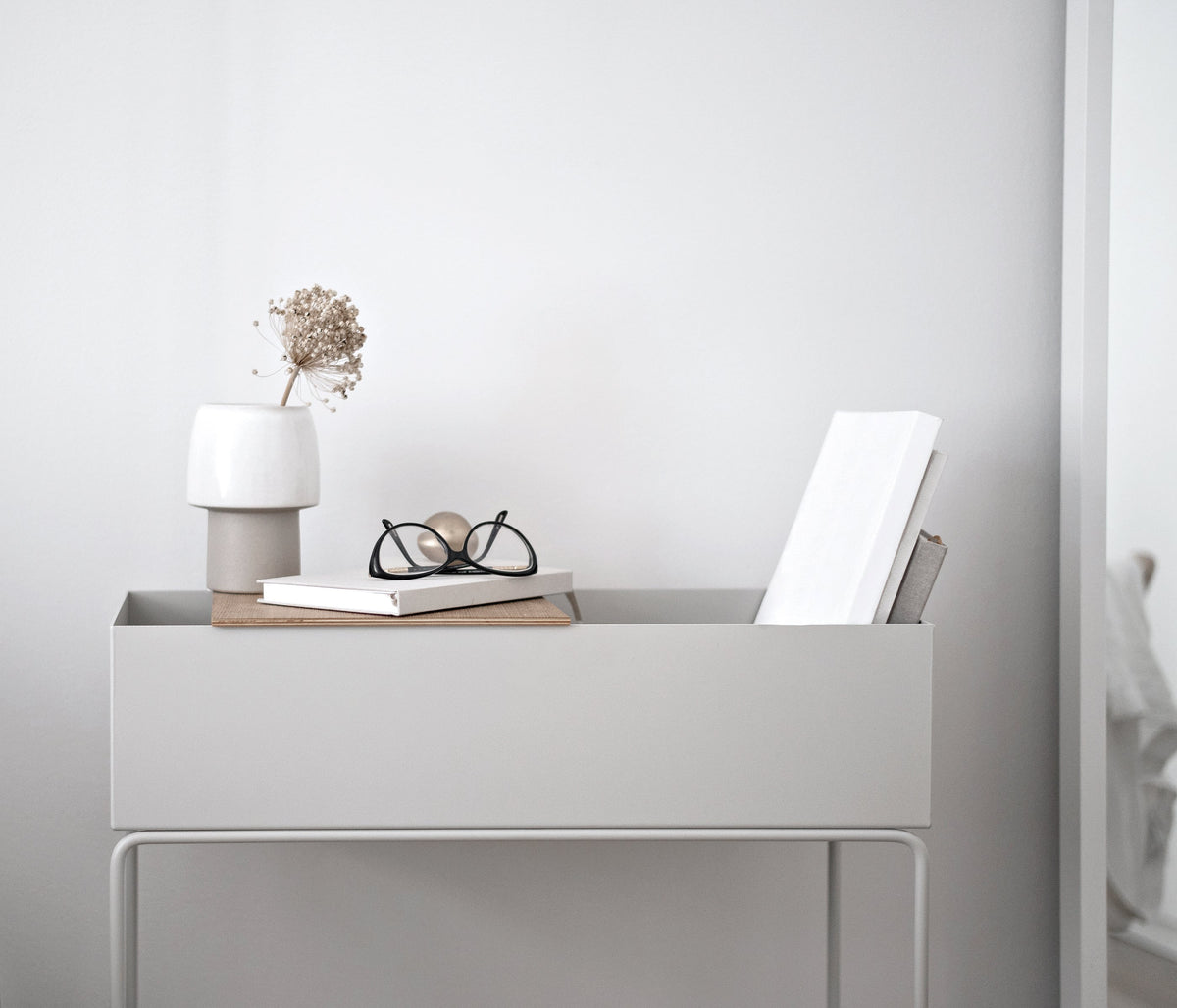 Grey metal planter box on legs holding glasses, books and a small lamp against a grey wall