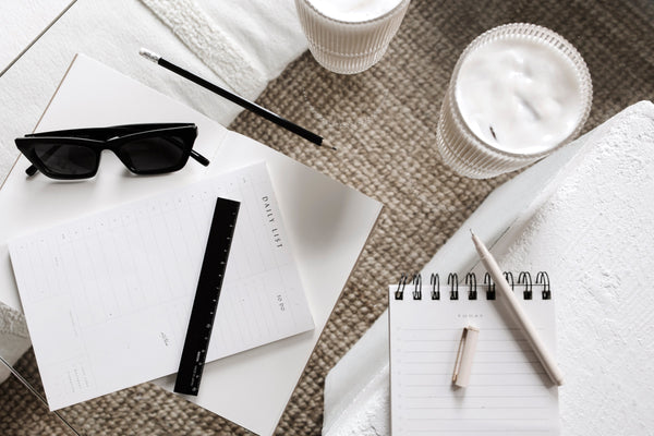 Flatlay of a minimalist workspace featuring a daily planner pad with a black ruler, a spiral notebook with pen, black cat-eye sunglasses, two white ribbed candles, and a black pencil on a textured beige rug and glass coffee table