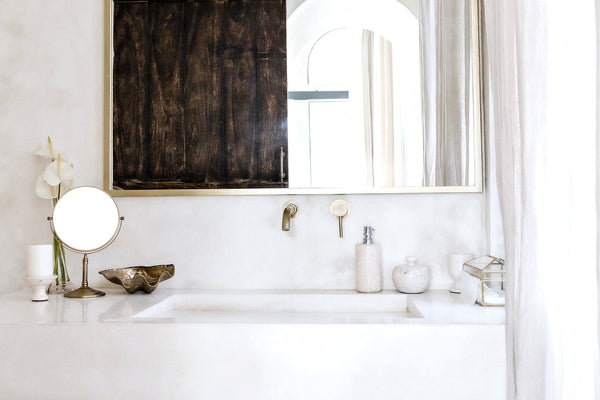 A serene bathroom vanity featuring a large framed mirror, brushed gold fixtures, minimalist decor, and soft natural light filtering through sheer curtains, creating a calming, spa-like atmosphere.