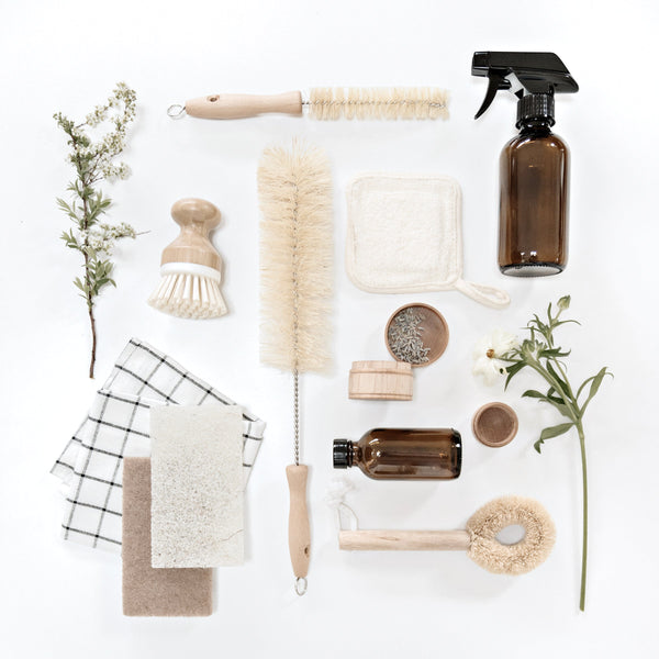 Flat lay of natural cleaning tools and accessories including wooden brushes, amber spray bottles, sponges, and dried flowers on a white background — styled to reflect calm, intentional living and clutter-free home design