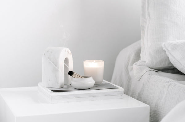 Minimalist bedside scene featuring a marble arch incense holder, a lit candle, and a small ceramic bowl resting on stacked books, styled alongside a cozy, textured bed for a serene and joyful daily ritual.