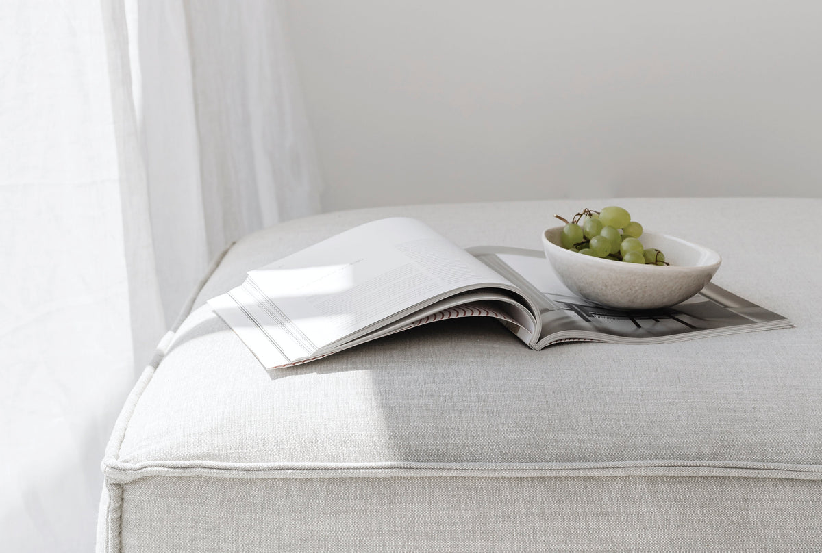 A cozy, sunlit scene featuring an open magazine and a small bowl of green grapes resting on a soft, upholstered ottoman, creating a calm and inviting atmosphere for relaxed reading and mindful living.