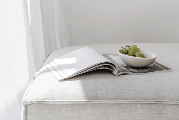 A cozy, sunlit scene featuring an open magazine and a small bowl of green grapes resting on a soft, upholstered ottoman, creating a calm and inviting atmosphere for relaxed reading and mindful living.