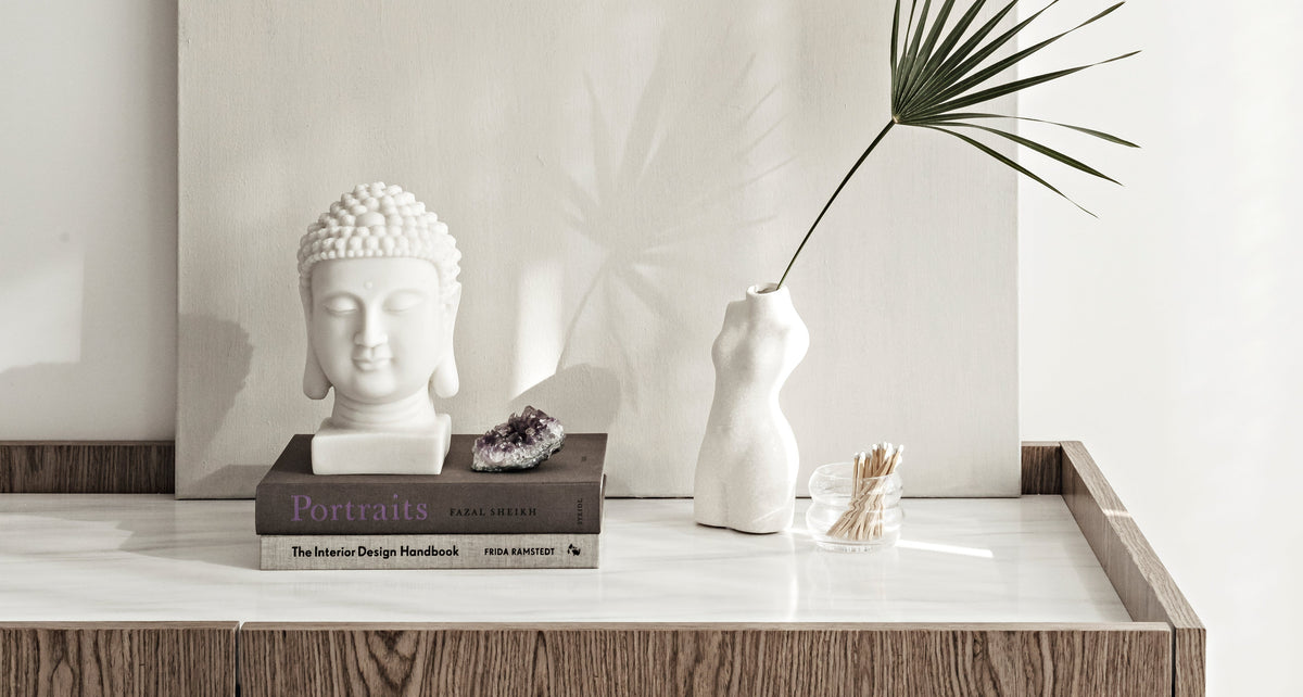 Serene tabletop vignette featuring a white Buddha head sculpture, minimalist torso vase with a single palm leaf, amethyst crystal, design books, and a jar of matches, styled on a marble and wood surface with soft natural light.