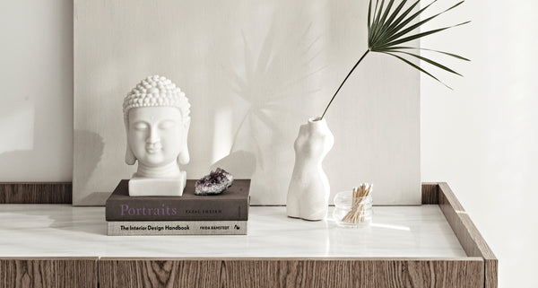 Serene tabletop vignette featuring a white Buddha head sculpture, minimalist torso vase with a single palm leaf, amethyst crystal, design books, and a jar of matches, styled on a marble and wood surface with soft natural light.