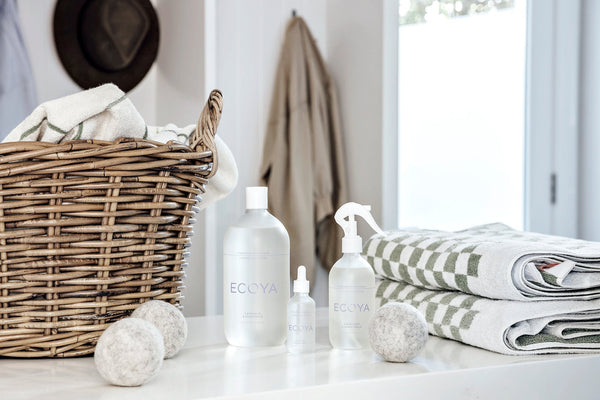 A calming laundry scene featuring a wicker basket filled with soft towels, ECOYA laundry products, wool dryer balls, and folded checkered towels, evoking a sense of fresh starts and nurturing rituals.