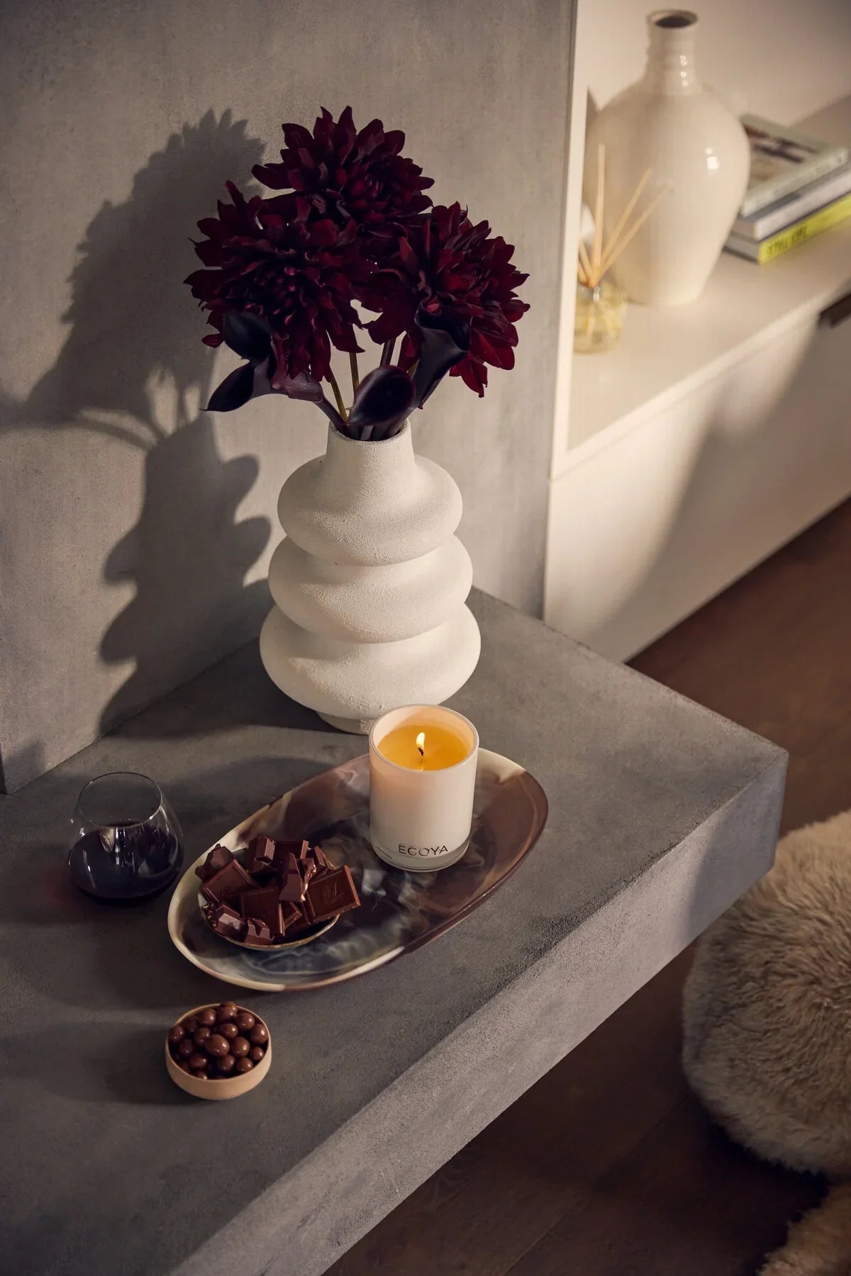 A lit Ecoya candle on a tray beside dark dahlias and chocolate on a concrete ledge at dusk