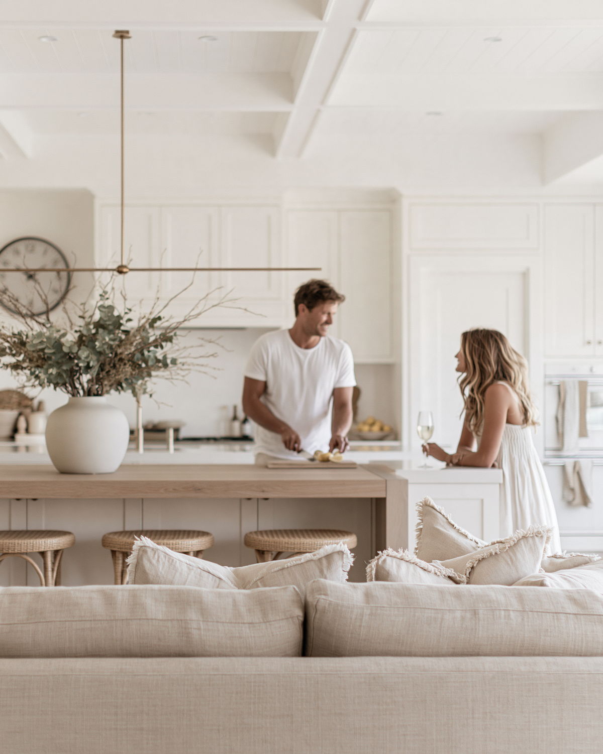 A couple chatting at a pale oak kitchen island in a white Nancy Meyers style kitchen with olive branches