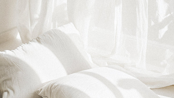 Soft morning light filters through sheer white curtains onto a bed with rumpled pillows, creating a serene, calming atmosphere that evokes rest and sensory ease.
