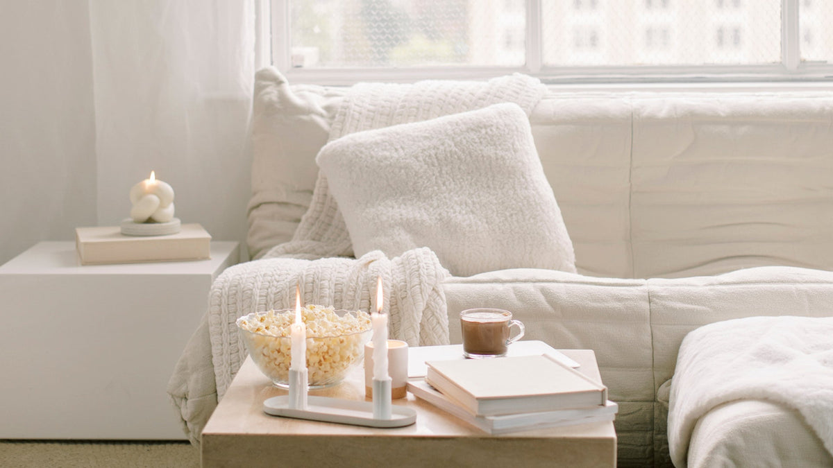 Cream boucle cushions and a waffle throw on a sofa, with lit taper candles and hot chocolate on a low white table