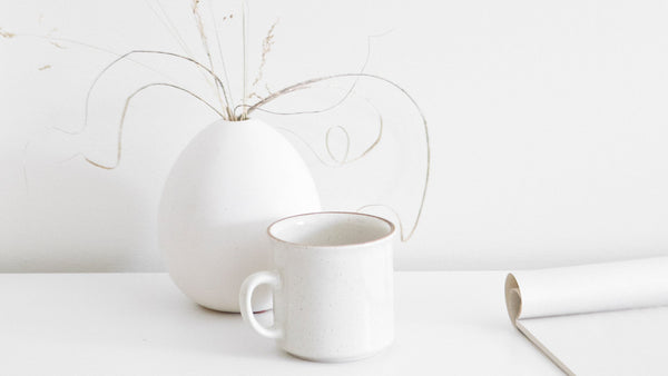 Minimalist white ceramic mug and vase with dried grasses on a white surface, styled with calming simplicity to evoke softness, stillness, and nervous system regulation.