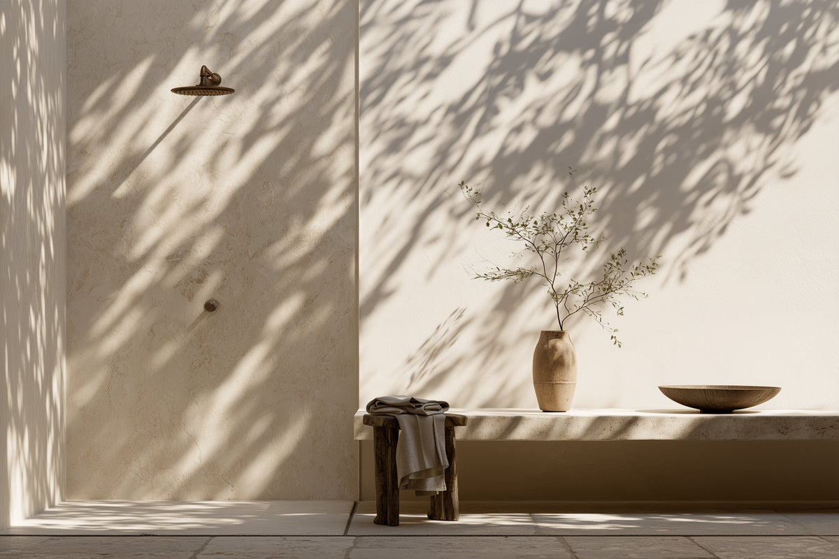 Travertine wall under dappled leaf shadows with a bronze rainfall shower, timber stool of linen towels and a stone ledge.