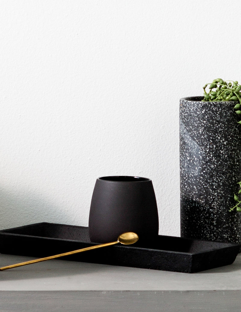 A Zakkia handmade brass statement spoon rests elegantly on a black tray alongside a food grade compliant potted plant.
