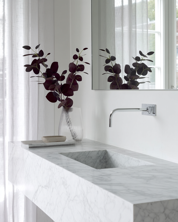 A tall ribbed glass vase holds deep burgundy eucalyptus stems on a marble vanity, with soft natural light filtering through sheer curtains for a moody, elegant atmosphere.