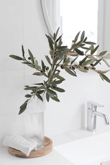 A tall faux olive branch with dark green leaves and small realistic olives, styled in a ribbed glass vase on a wooden tray in a bright white bathroom.