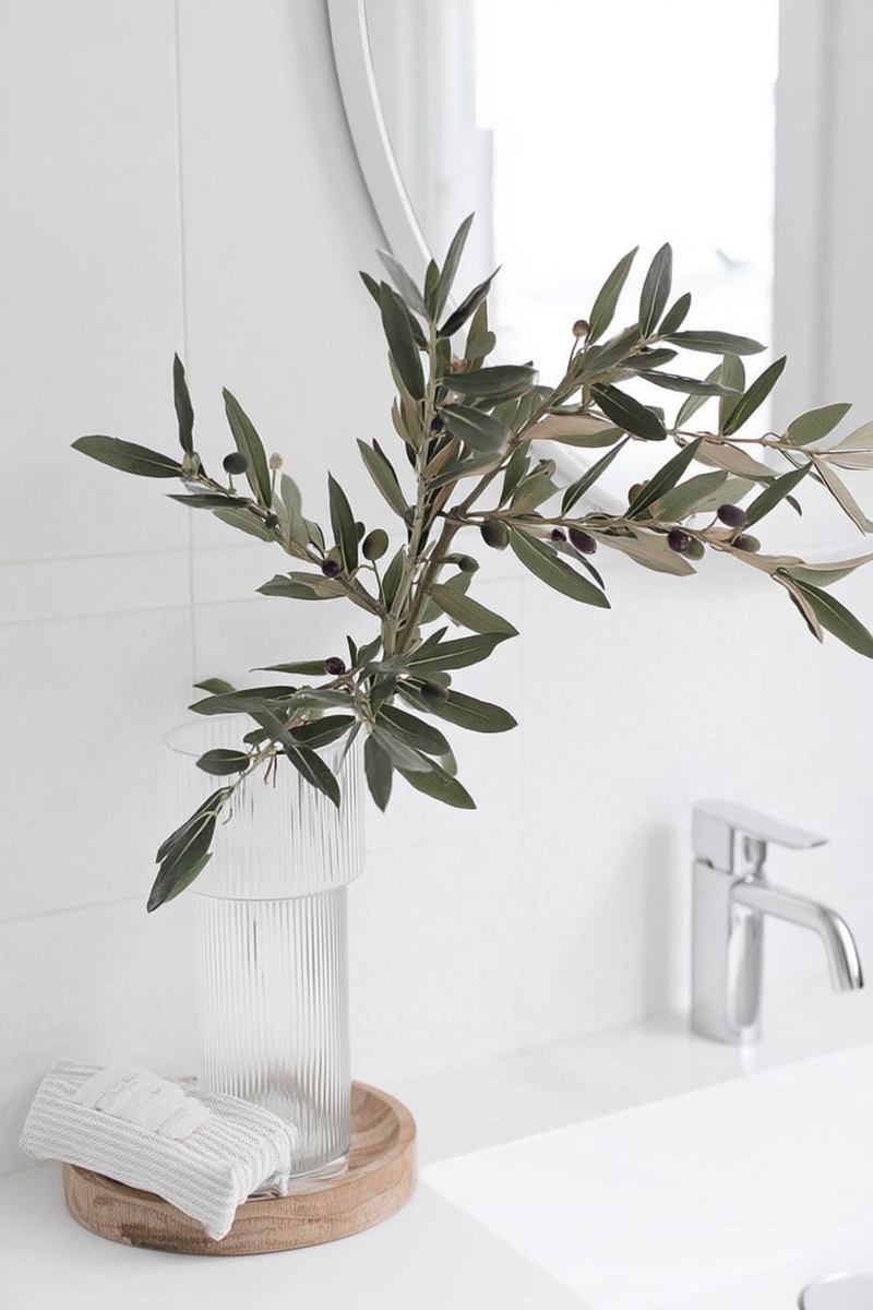 A tall faux olive branch with dark green leaves and small realistic olives, styled in a ribbed glass vase on a wooden tray in a bright white bathroom.