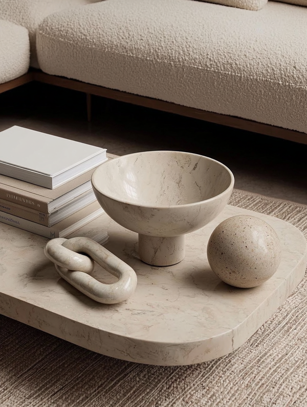 Minimalist coffee table styling featuring a solid white marble loop sculpture, marble pedestal bowl, stone sphere, and stacked neutral books beside a cream boucle sofa.