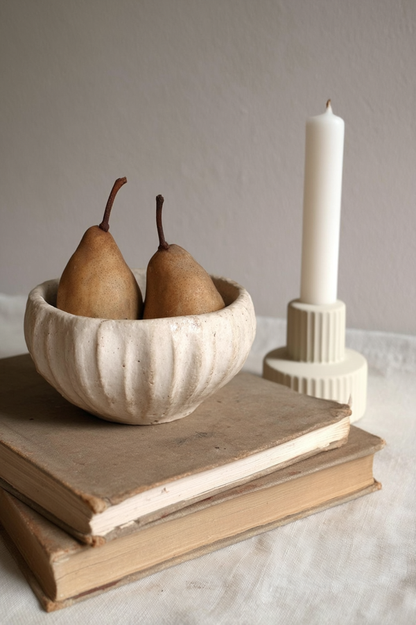 A ridged ceramic candle holder styled with a white taper candle, paired with a rustic bowl of pears and vintage books, creating a warm, sculptural vignette on a linen-covered surface.
