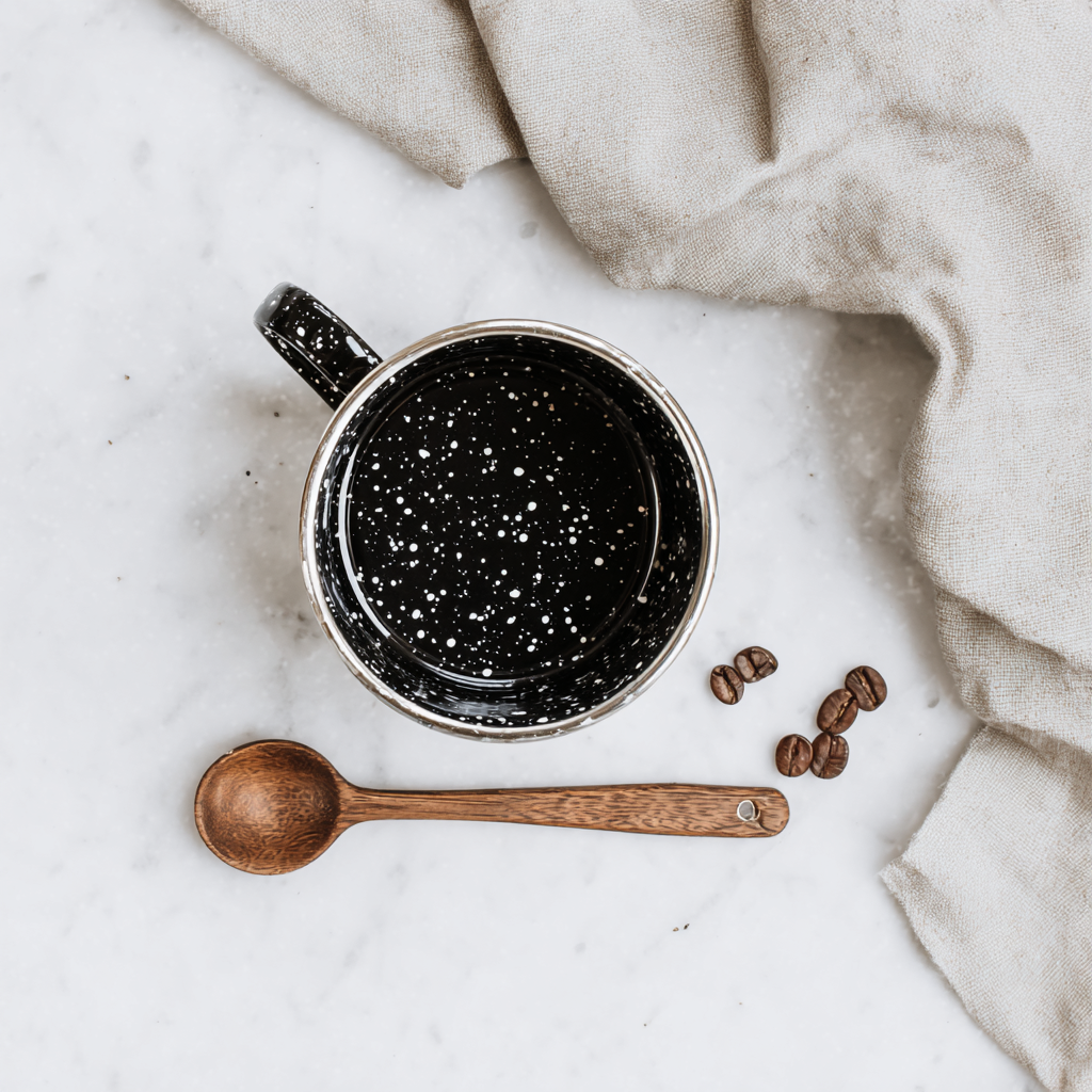 Overhead of the speckled enamel mug of dark coffee on marble, with a wooden spoon and coffee beans