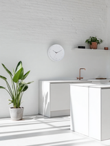Minimalist white wall clock with black markings in a bright, sunlit kitchen featuring white cabinetry, a copper tap, and potted plants against a white brick wall.
