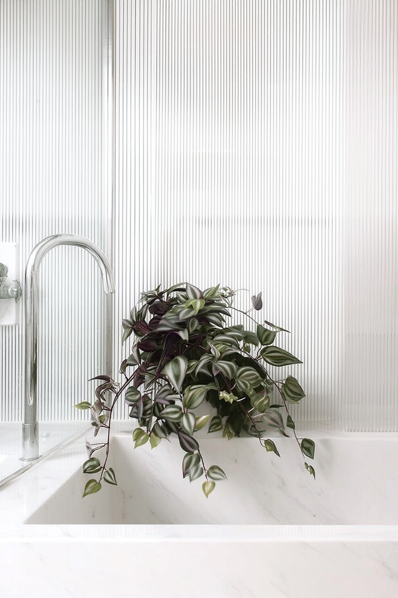 A realistic artificial trailing plant with variegated green and purple leaves styled in a modern white sink with vertical ribbed glass behind.