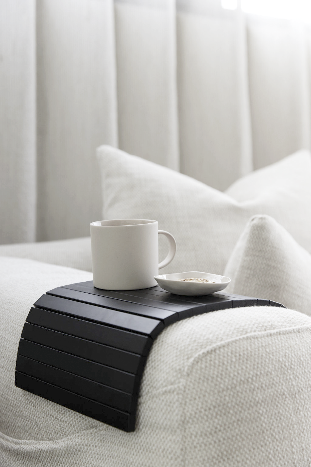 Black slatted armchair tray draped over a white sofa arm holding a ceramic mug and small plate