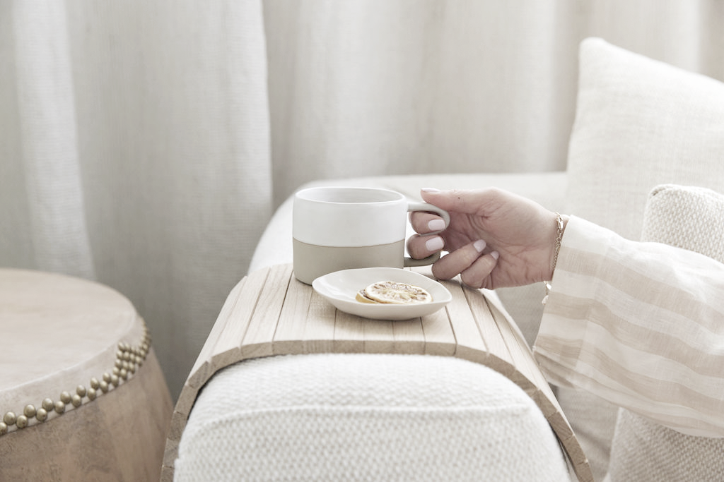 Hand lifting a mug from the oak slatted armchair tray on a pale sofa arm, beside a plate of dried lemon