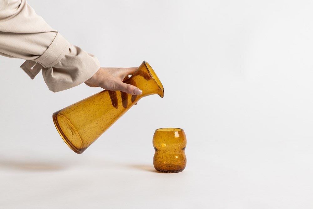 Hand pouring from a carafe beside a tumbler, both in a deeper amber not sold on this listing
