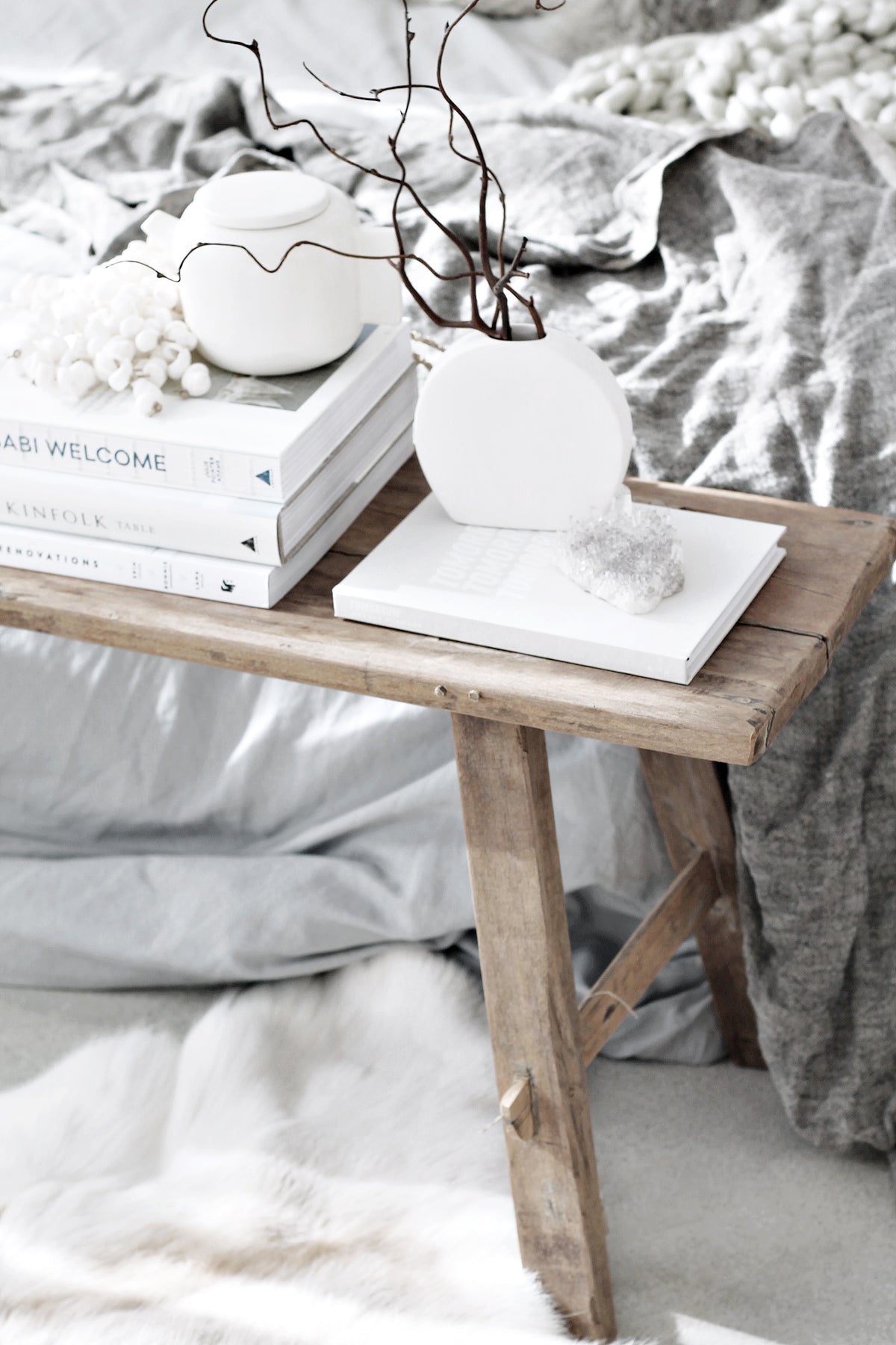 Teak bench holding a round white vase of bare branches and books beside a bed