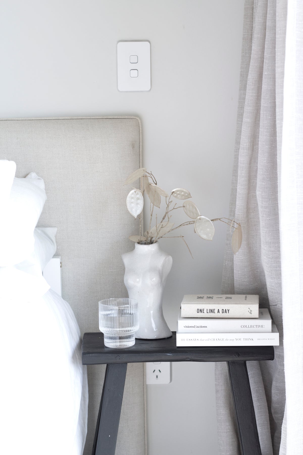 Black bench beside an upholstered bed head, holding a torso vase of dried stems, books and a water glass