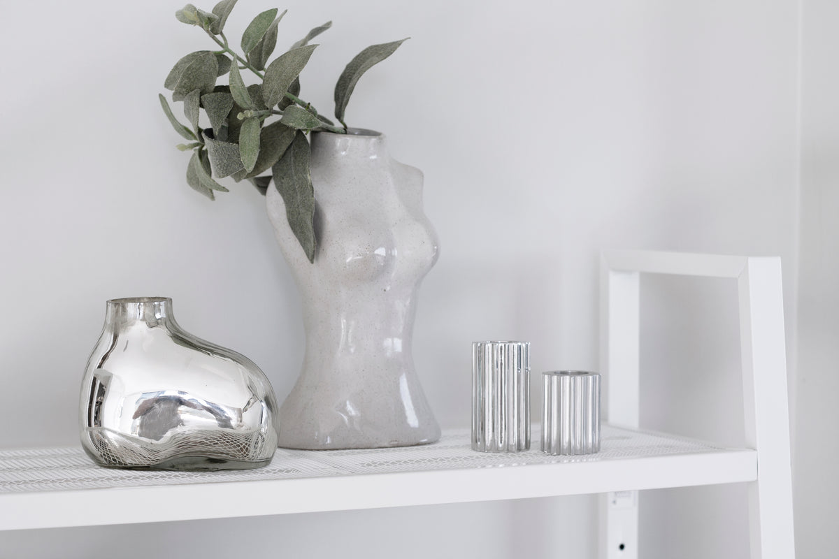 Female Silhouette Vase with foliage on a white shelf beside a silver vase and glasses