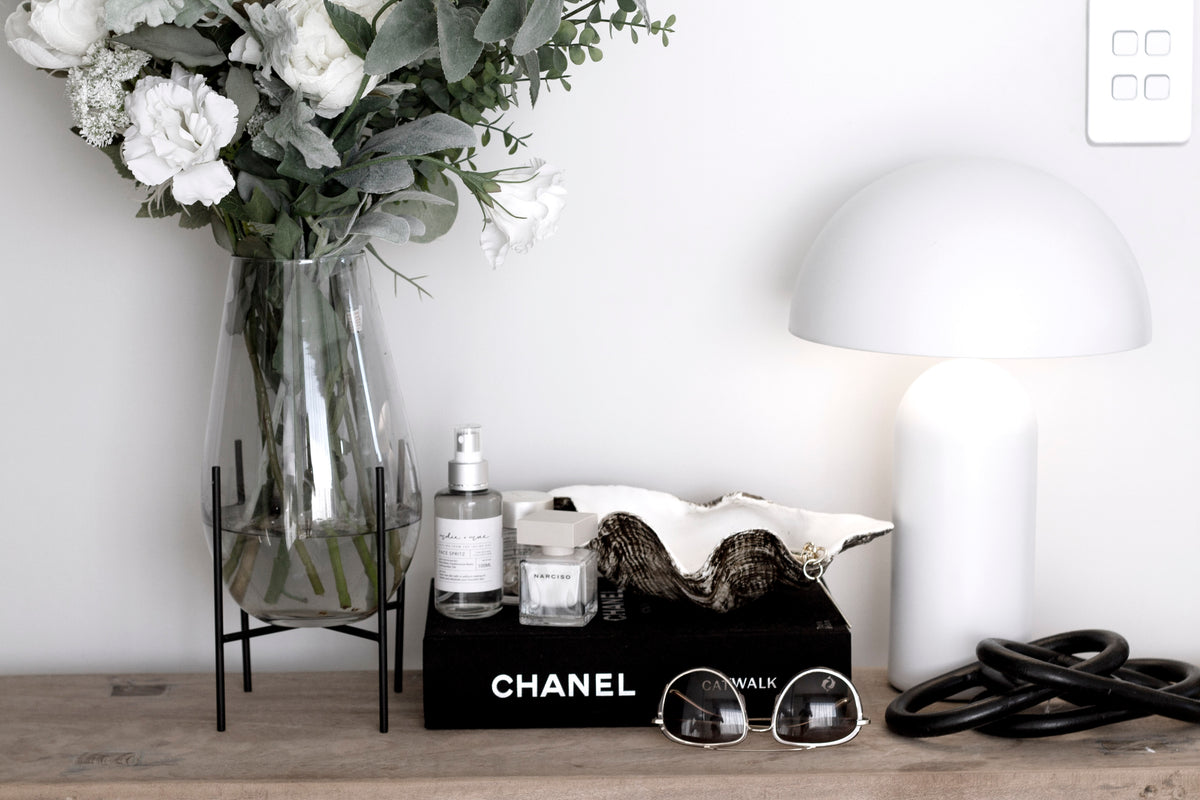 Chanel Catwalk book on a timber shelf beside a mushroom lamp, perfume and a vase of white flowers