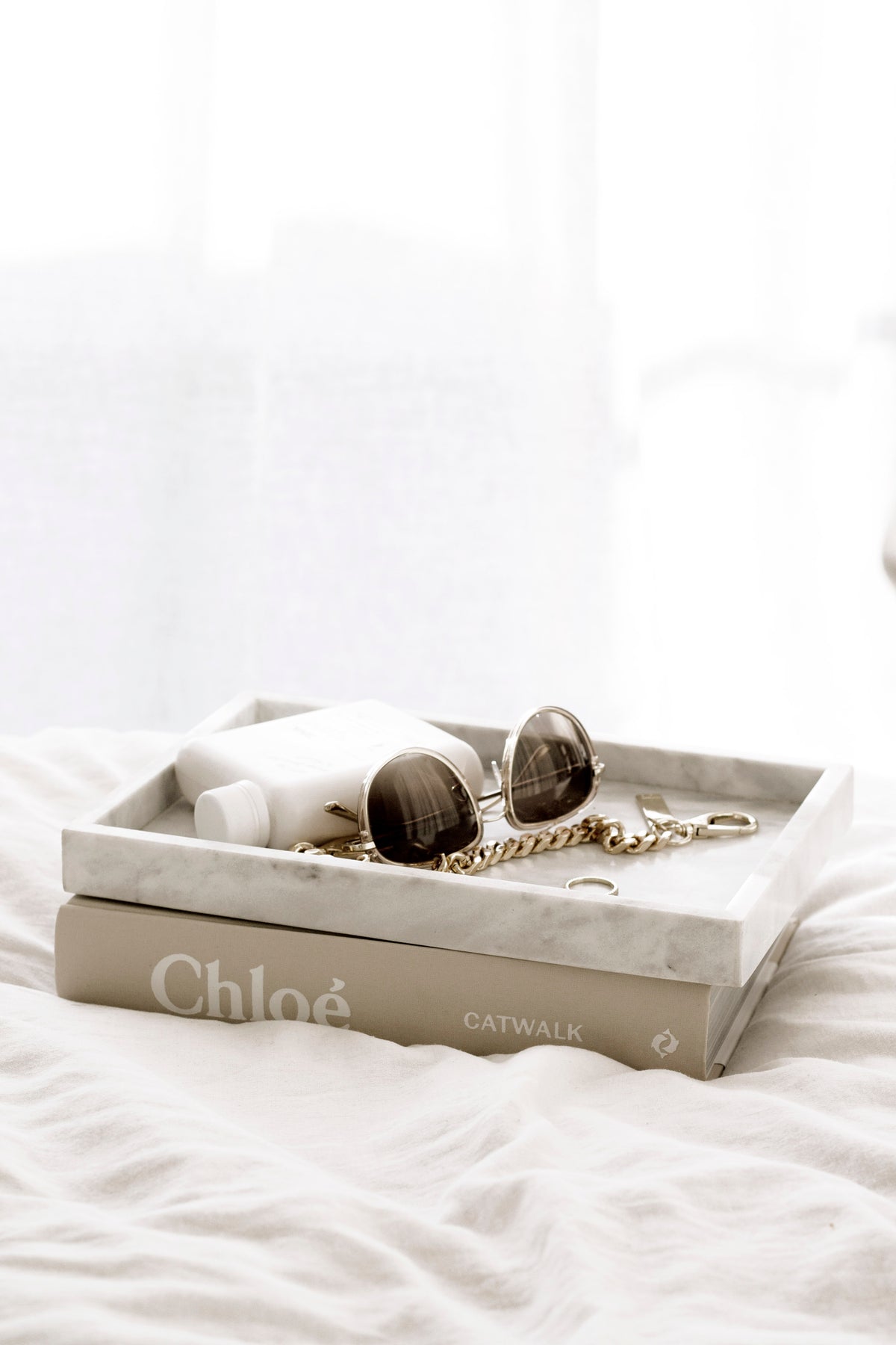 Grey marble tray on white bedding holding sunglasses and small accessories on a stack of books