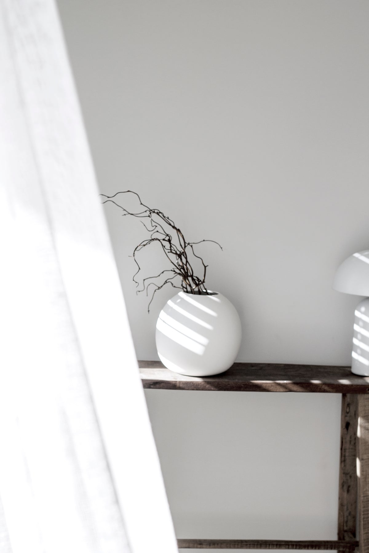 Recycled teak console with a round white vase of twigs and a mushroom lamp by a sheer curtain