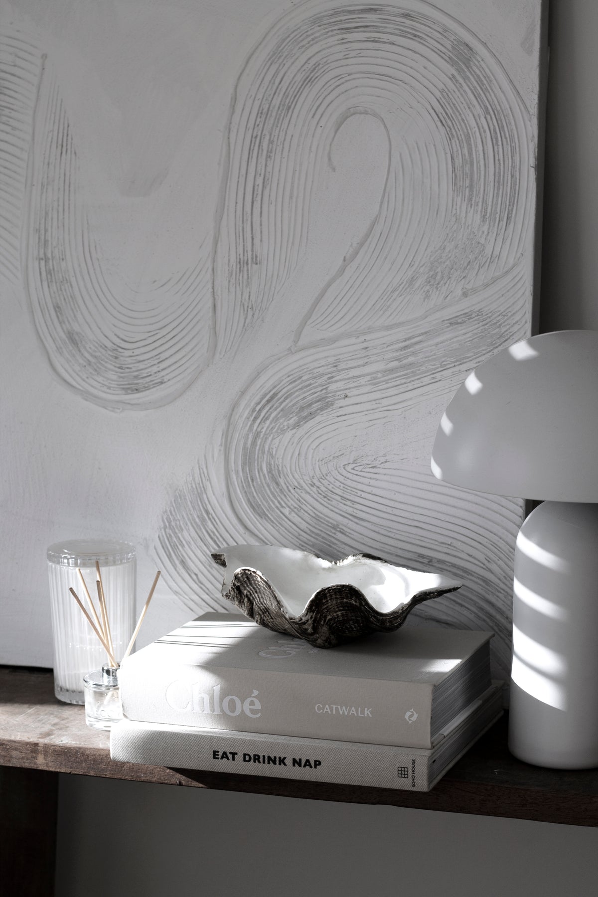 Clam shell dish on stacked books with a reed diffuser and a dome lamp, abstract artwork behind