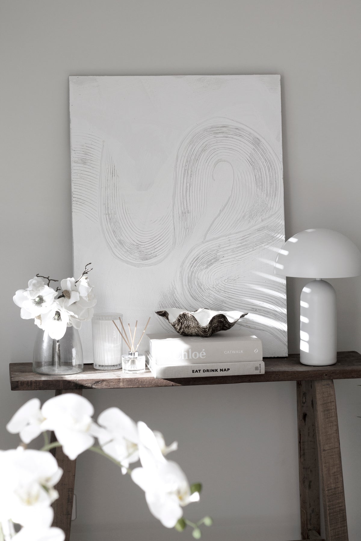 Clam shell dish on a timber console with white orchids, a dome lamp and an abstract line artwork