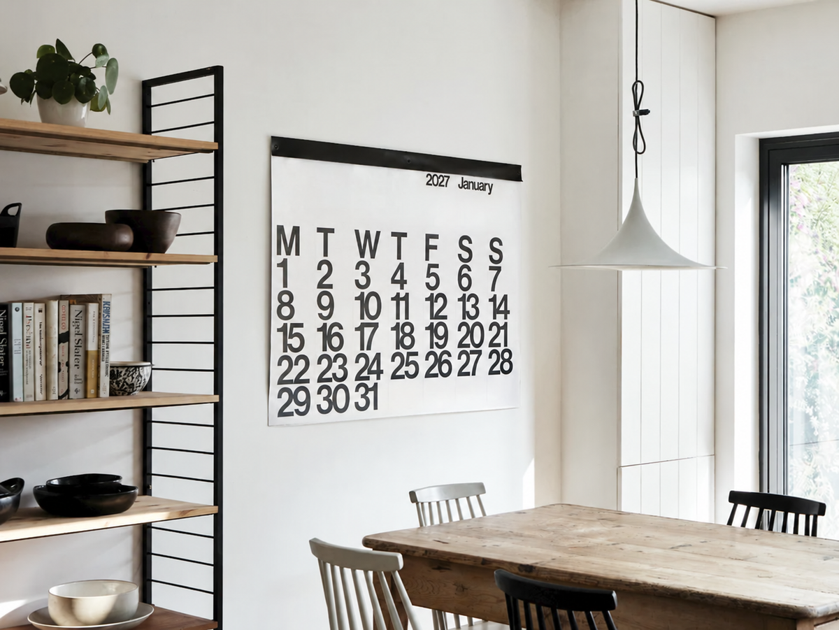 2027 Stendig Calendar hung on a white dining room wall beside black wall shelving with timber shelves, books and a plant, above a timber dining table with black chairs