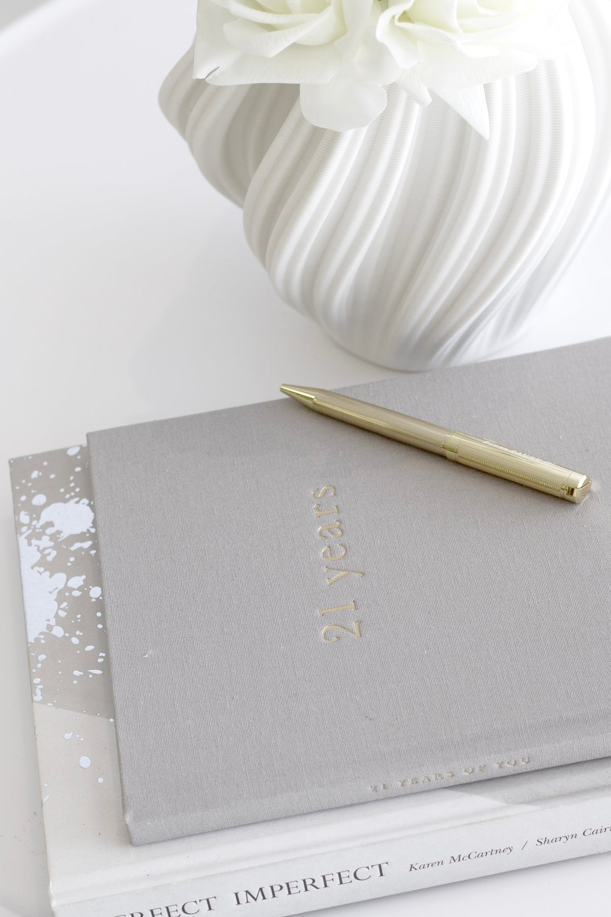 Grey linen 21 Years journal with a gold pen beside a white sculptural vase
