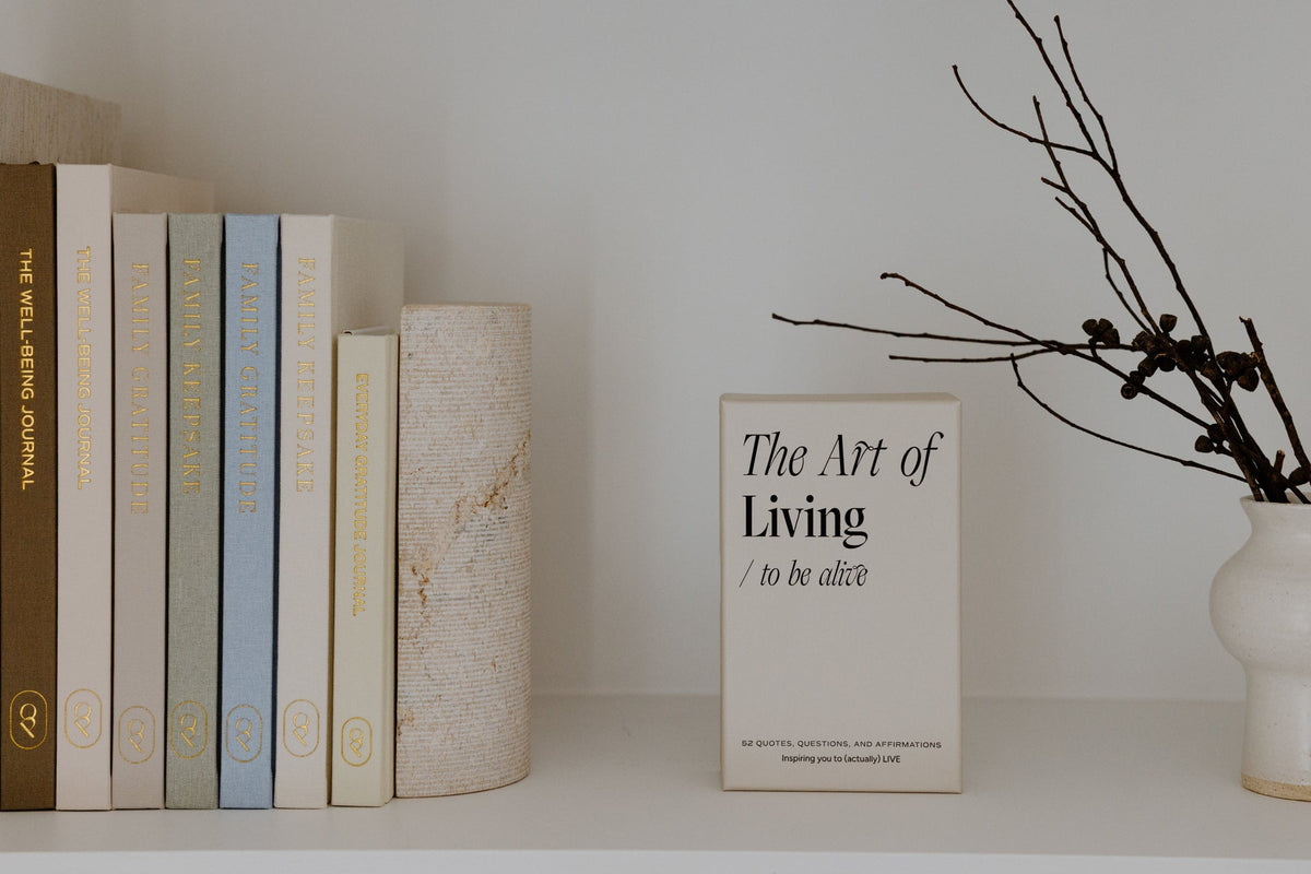 The Art of Living card deck box on a white shelf beside a row of books and a vase of bare branches