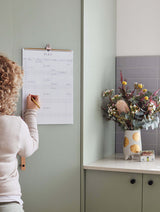 A girl managing family chaos by writing on a calendar in a kitchen is using the Write To Me 2024 FAMILY CHAOS PLANNER.