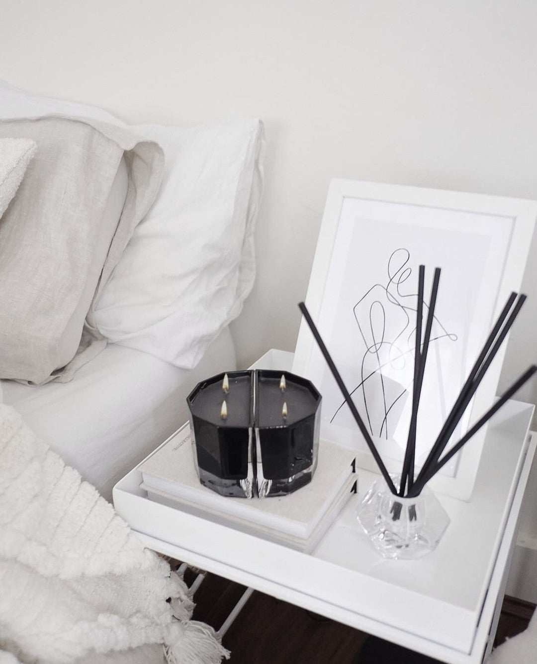 Blend candle pair on a white bedside table beside a black reed diffuser and a line drawing print