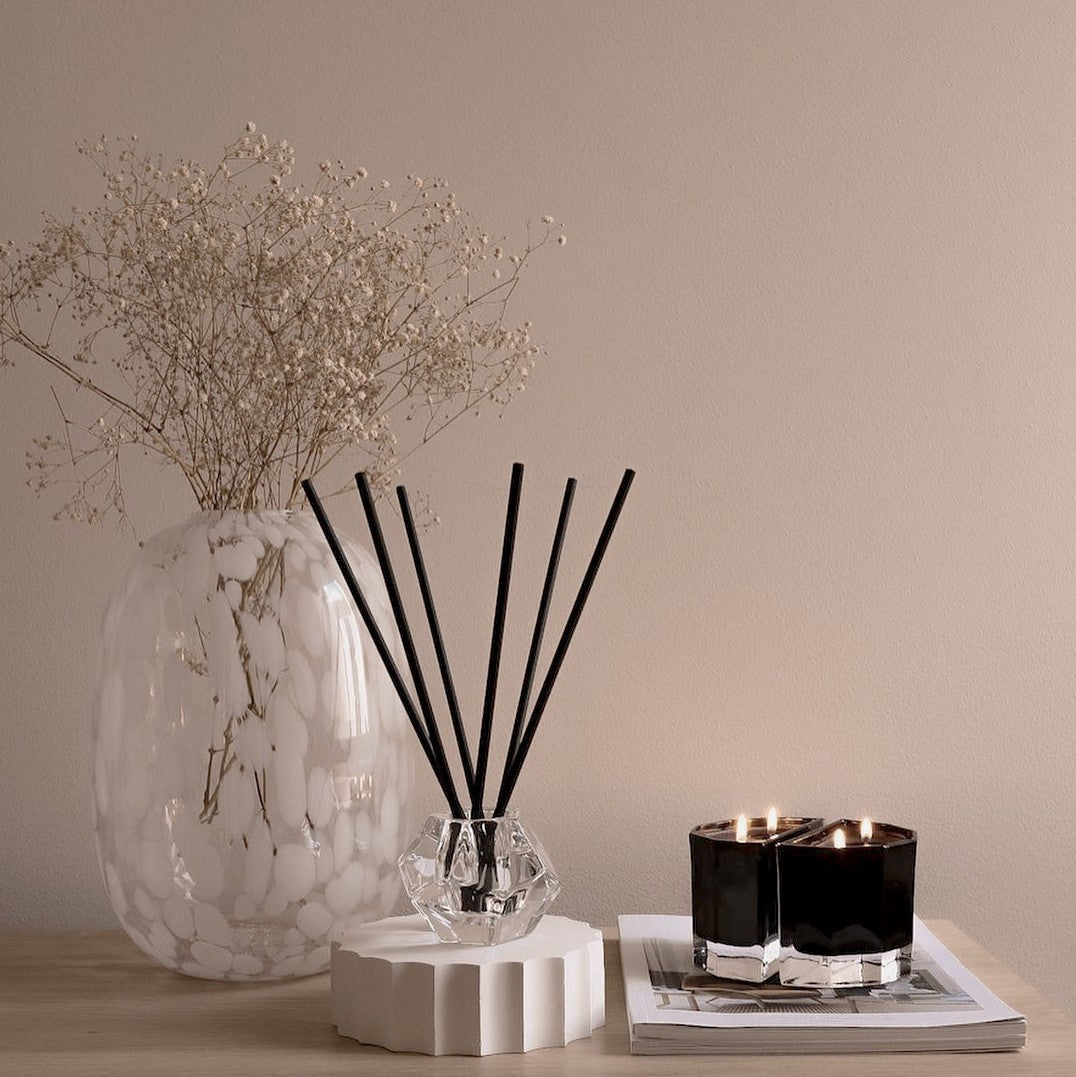 Aroma stick holder beside a speckled glass vase of dried flowers and two lit black candles on a console