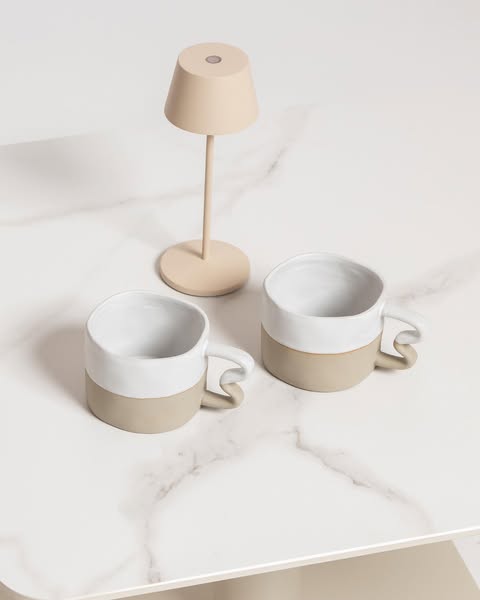 Two modern ceramic mugs in neutral tones with sculptural handles styled on a white marble tabletop beside a small minimalist beige table lamp.