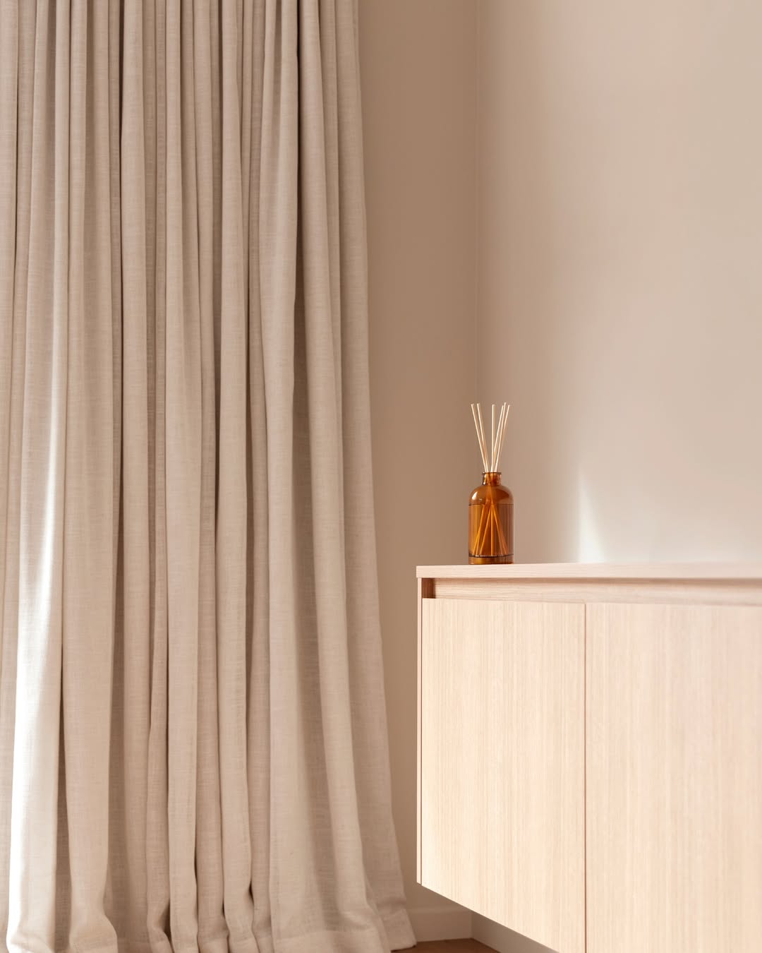 Amber reed diffuser on a pale oak sideboard beside floor-length linen curtains