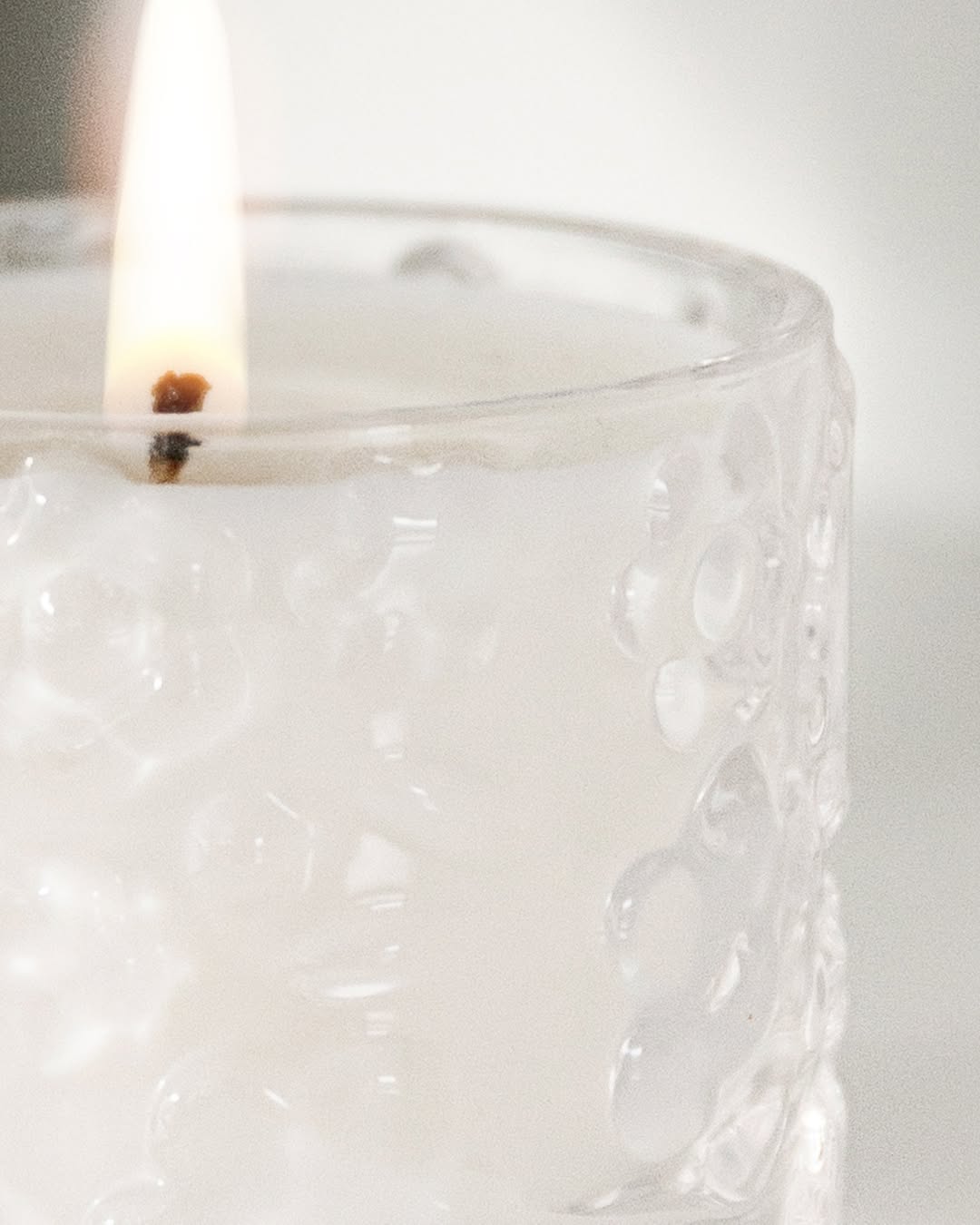 Close view of a lit FLWR candle, the flame above white wax and raised floral embossing on the glass