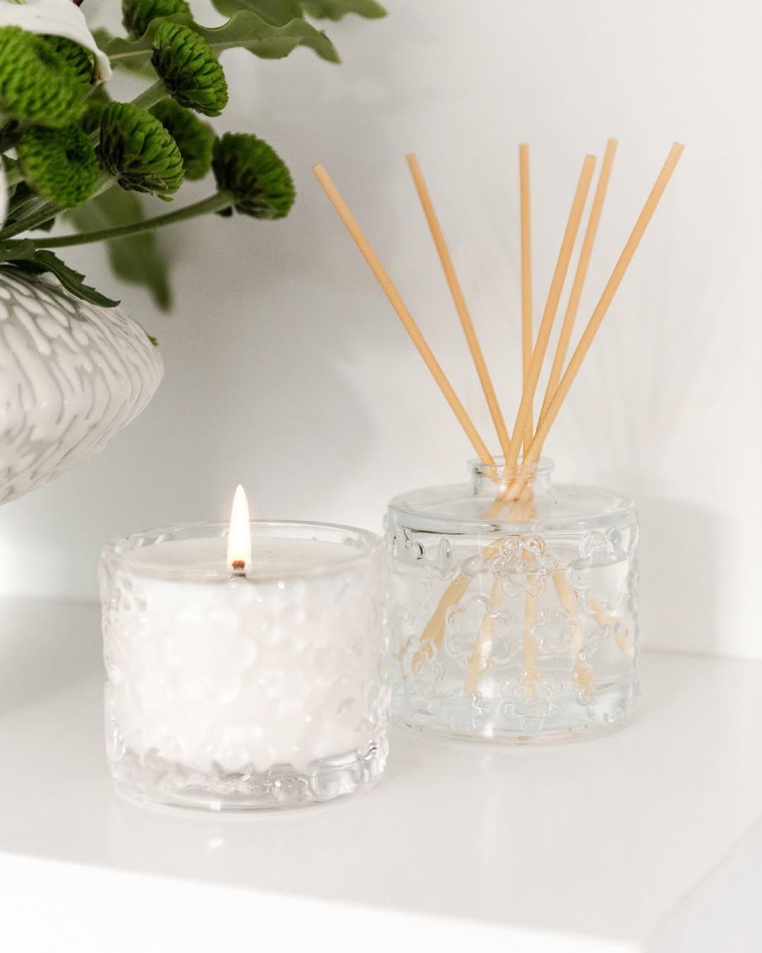 A lit FLWR candle in embossed glass beside a reed diffuser on a white ledge, greenery behind