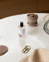 ECOYA Sweet Pea & Jasmine room spray on a round stone table, styled with a terracotta pot plant, straw hat, and brass keyring in a relaxed, sunlit setting.