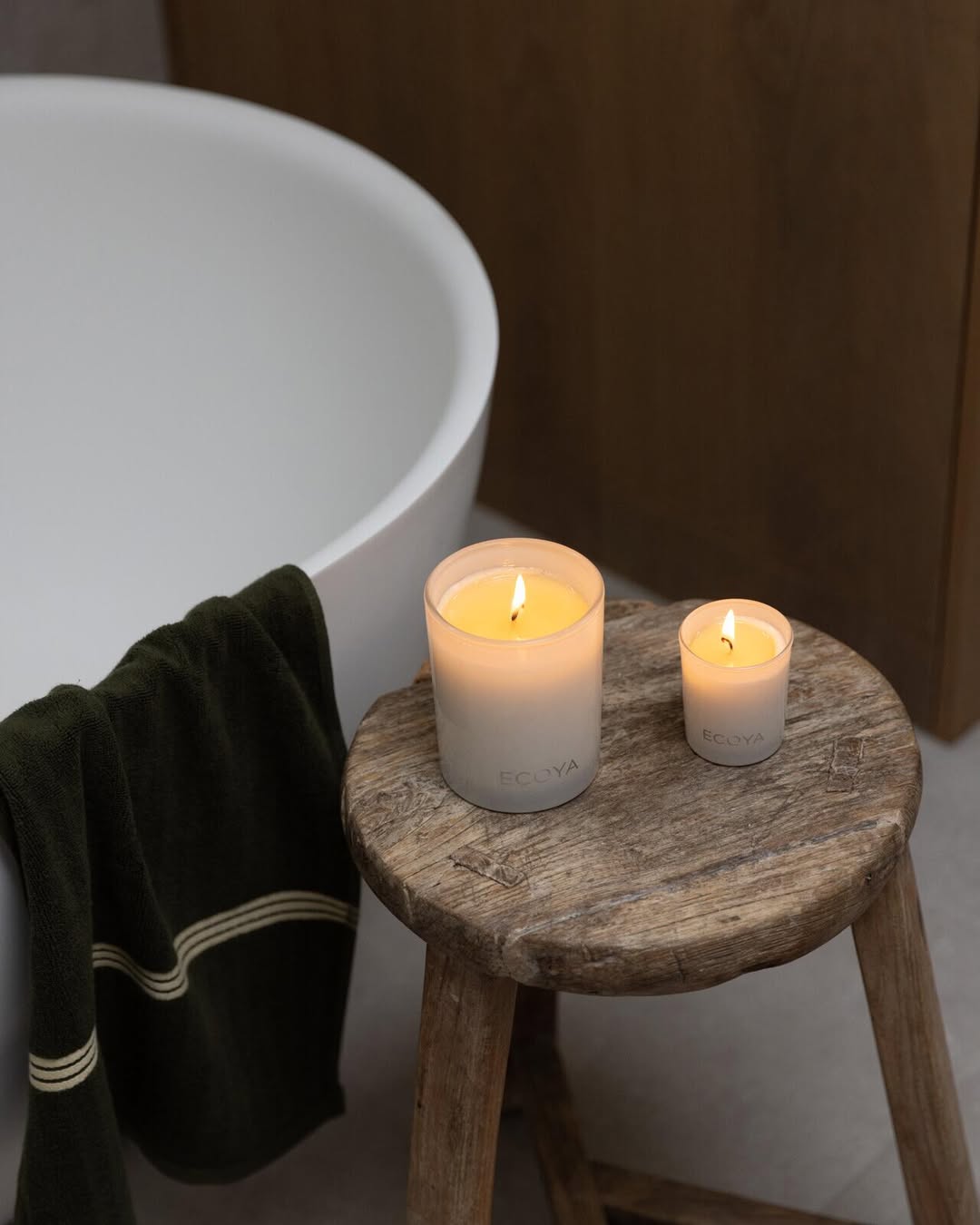 Two lit Ecoya Madison candles on a timber stool beside a freestanding bath
