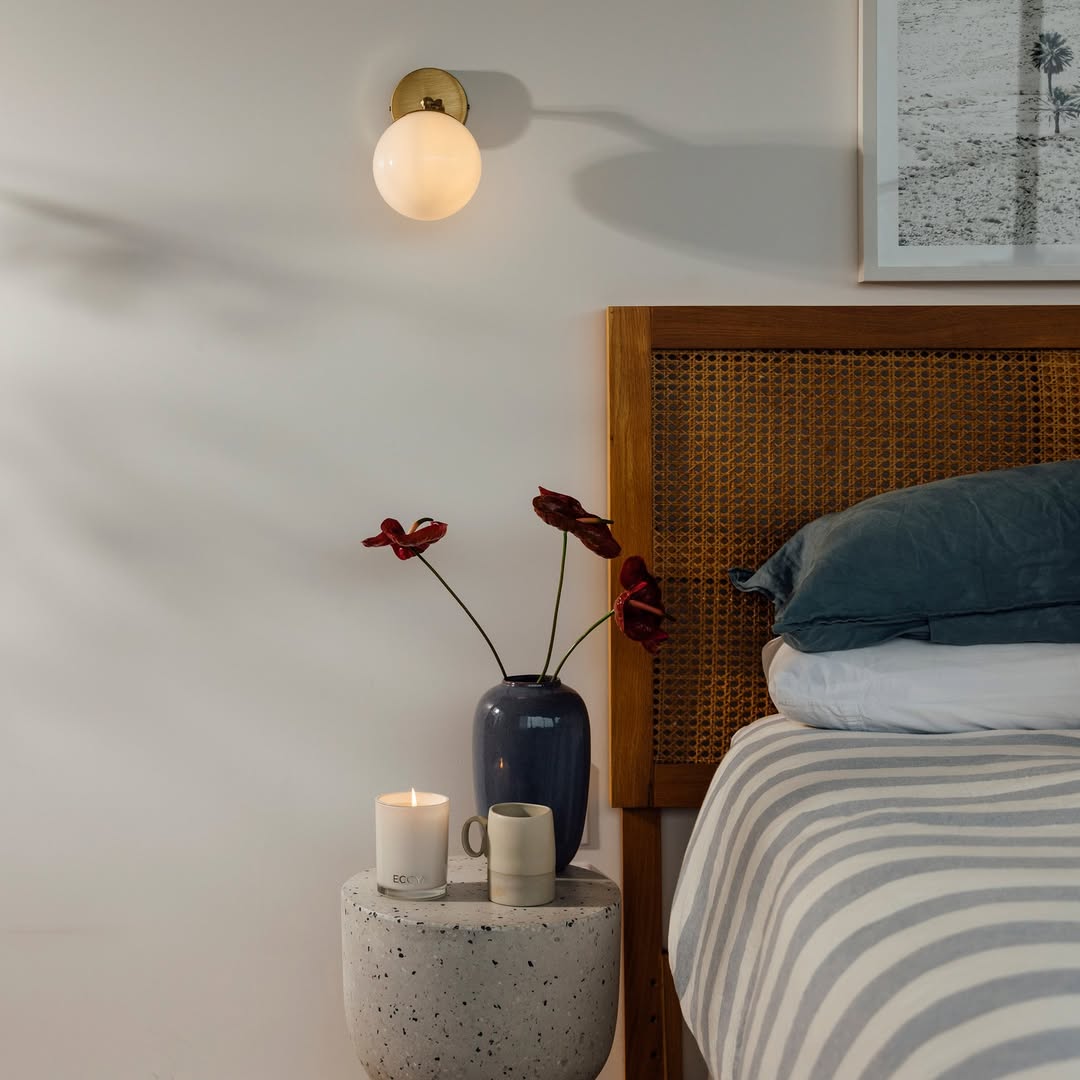 A lit Ecoya Madison candle and a mini candle on a terrazzo bedside table beside a vase of anthuriums, next to a rattan bed